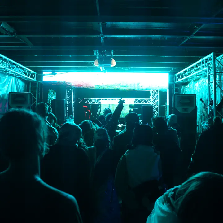 The crowd from behind at Cosmic Convergence, lit aqua, facing the DJ booth and a glowing screen