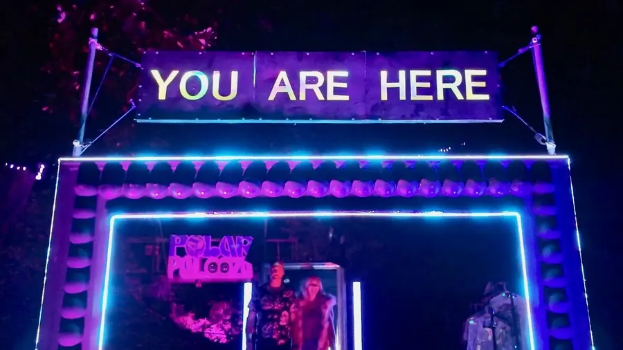 The runway art piece at night: a neon-lit frame under a YOU ARE HERE sign, two people walking out